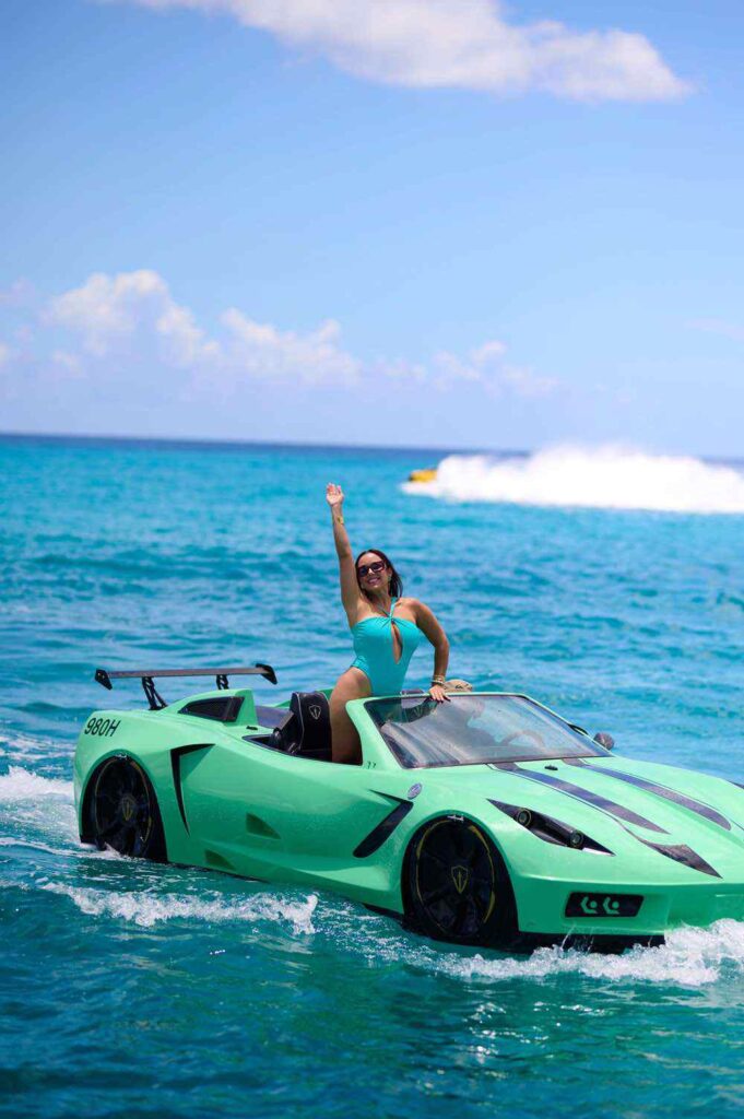 Woman in teal swimsuit waving from green sports car on water